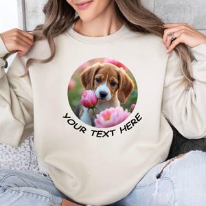 May include: A white sweatshirt with a round graphic of a beagle puppy sniffing a pink flower. The text "YOUR TEXT HERE" is printed below the graphic.