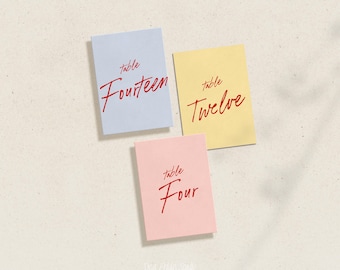 May include: Three table number cards with red handwritten text on different colored backgrounds. The cards read "table fourteen", "table twelve", and "table four".