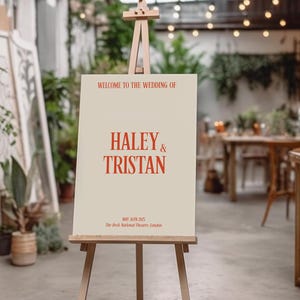 May include: A white easel with a sign announcing the wedding of Haley & Tristan. The sign is on a beige background with red text. The text reads "Welcome to the wedding of Haley & Tristan, May 30th 2023, The Deck National Theatre, London."