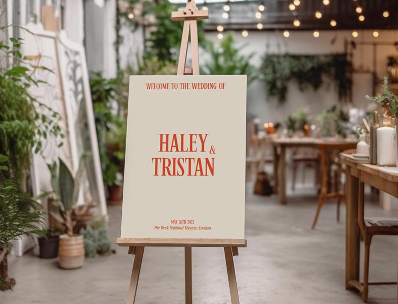 May include: A white easel with a sign announcing the wedding of Haley & Tristan. The sign is on a beige background with red text. The text reads "Welcome to the wedding of Haley & Tristan, May 30th 2023, The Deck National Theatre, London."