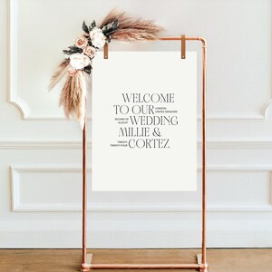 May include: A white sign with a copper stand, decorated with dried pampas grass and flowers. The sign reads "Welcome to our wedding Millie & Cortez, Second of August, Twenty Four, London, United Kingdom".