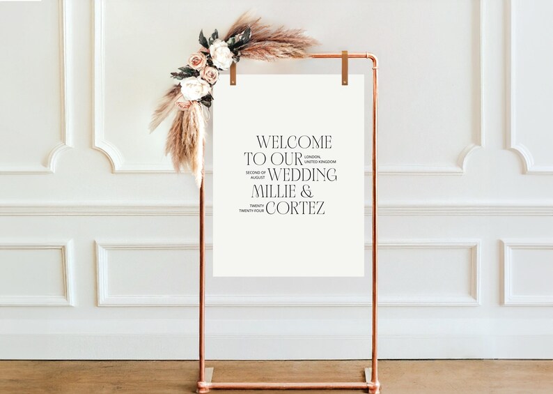 May include: A white sign with a copper stand, decorated with dried pampas grass and flowers. The sign reads "Welcome to our wedding Millie & Cortez, Second of August, Twenty Four, London, United Kingdom".
