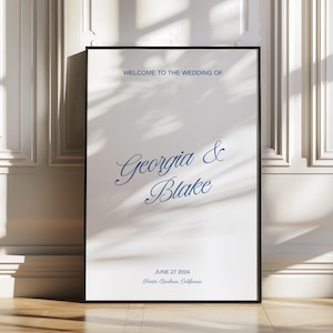 May include: A framed wedding sign with the text "WELCOME TO THE WEDDING OF Georgia & Blake" in navy blue script. The date "JUNE 27 2024" and location are also displayed. The sign is displayed on a wooden floor.