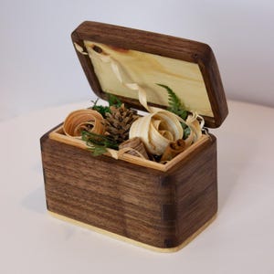 May include: A small wooden box with a hinged lid, open to reveal a variety of natural materials, including pine cones, wood shavings, cinnamon sticks, and green foliage.