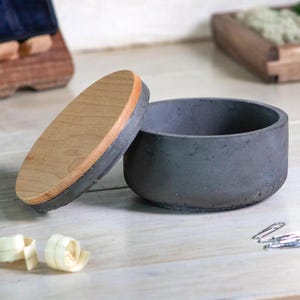 May include: A gray concrete bowl with a wooden lid. The lid is round with a light wood center and a dark gray edge. The bowl is open, revealing a dark interior. The items are on a light wood surface.