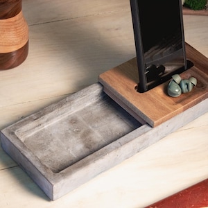 May include: A wooden phone stand with a slot for a phone and a small tray for earbuds. The stand is made of light brown wood and sits on a gray concrete base. The phone is black.