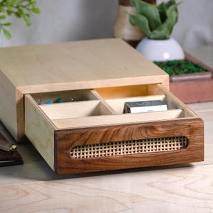 May include: A wooden jewelry box with a light-colored top and a dark brown drawer. The drawer has a woven pattern and is partially open, revealing compartments. The box is on a wooden surface.