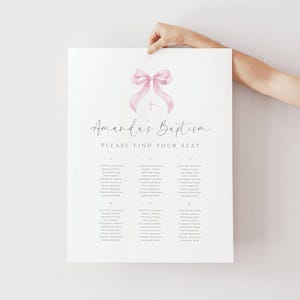 Puede incluir: Un plano de asientos blanco para el bautizo de Amanda, sostenido por una persona. Presenta un lazo rosa y un diseño de cruz en la parte superior. El texto dice "Amanda's Baptism" y "Please Find Your Seat". Los nombres están listados en columnas.