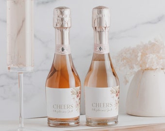 Op de afbeelding: Twee kleine flessen mousserende wijn met aangepaste etiketten waarop "Cheers to the forever us" staat. De etiketten hebben een bloemmotief met roze en gouden accenten. De flessen staan op een houten oppervlak met een witte achtergrond.