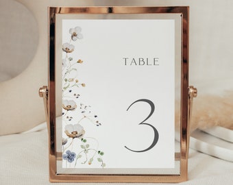 May include: A white table number card with a floral design in shades of blue and yellow. The card is in a gold frame and the number "3" is printed in grey. The text "TABLE" is printed above the number.