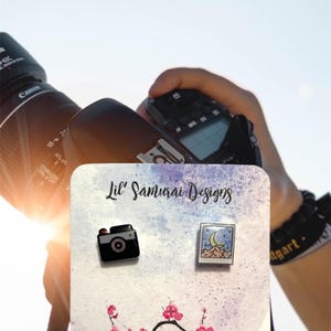 May include: Two enamel pins on a card with the text "Lil' Samurai Designs". One pin is a black camera, the other a landscape with a moon. A branch with pink flowers is at the bottom.