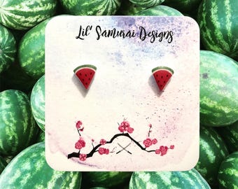 Könnte beinhalten: Ohrringe in Form von Wassermelonenscheiben auf einer dekorativen Karte mit dem Text "Lil' Samurai Designs". Die Ohrringe sind rot, grün und schwarz. Die Karte hat einen Aquarellhintergrund mit einem Blumen- und Schwertdesign. Der Hintergrund ist ein Haufen Wassermelonen.