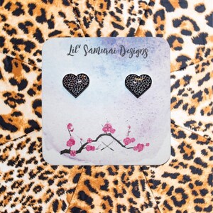 May include: A pair of heart-shaped stud earrings with a leopard print design. The earrings are displayed on a card with the text "Lil' Samurai Designs" and a floral illustration. The background features a leopard print pattern.