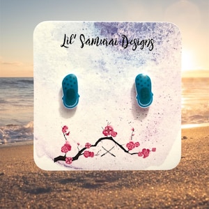 May include: Teal clog-shaped earrings on a white card with the text "Lil' Samurai Designs". The card has a watercolor design with a floral branch and a beach scene background.