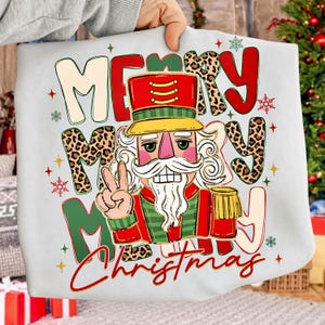 Può includere: Una felpa bianca con la grafica di uno schiaccianoci in uniforme rossa e verde che fa il segno di vittoria. Il testo "MERRY MERRY CHRISTMAS" è stampato in grandi lettere colorate e con stampa leopardata, accentuato da piccoli fiocchi di neve rossi e verdi.