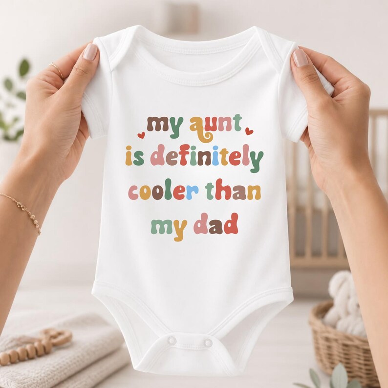 May include: A white short-sleeved baby bodysuit featuring the colorful text "my aunt is definitely cooler than my dad" in a retro font, accented with small red hearts. The garment has a round neckline and snap closures at the bottom.