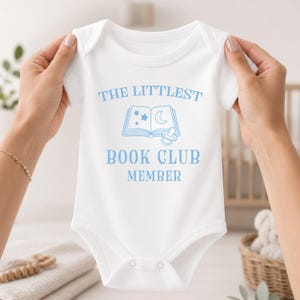 May include: A white short-sleeved baby bodysuit featuring light blue text that reads THE LITTLEST BOOK CLUB MEMBER, accompanied by a graphic of an open book with stars, a crescent moon, and a pacifier illustration centered on the chest.