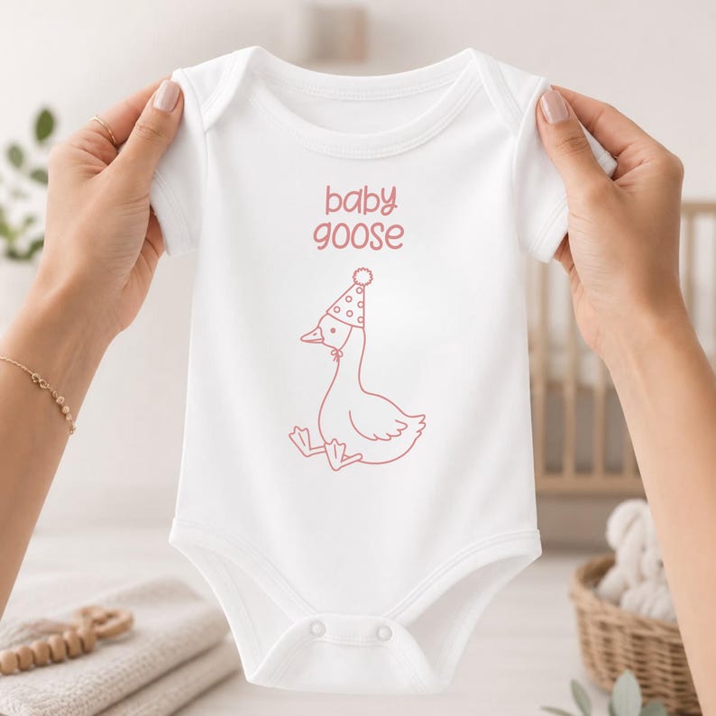 May include: A white short-sleeved baby bodysuit featuring a pink line-art illustration of a goose wearing a polka-dot party hat. The text "baby goose" is printed in pink above the goose graphic. The garment has a crew neckline and snap closures.