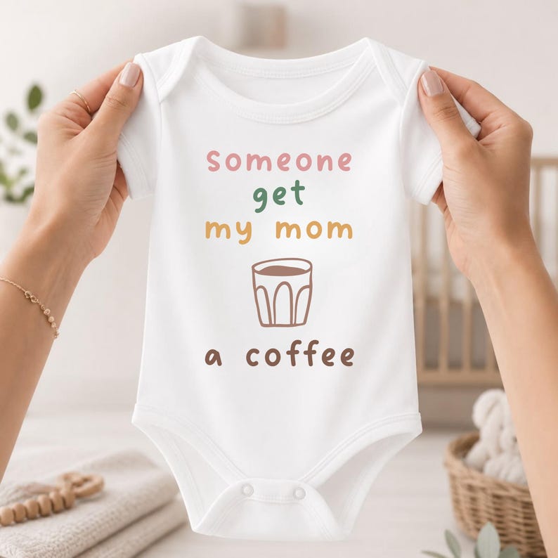 May include: A white short-sleeved baby bodysuit featuring the text "Someone get my mom a coffee" in multi-colored lettering, with a brown line-art illustration of a coffee cup centered between the words "my mom" and "a coffee".