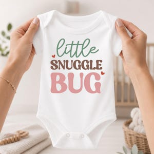 May include: A white short-sleeve baby bodysuit featuring the text little SNUGGLE BUG in green, brown, and pink lettering. The word little is in a cursive green font, SNUGGLE is in a brown sans-serif font, and BUG is in a large pink font.