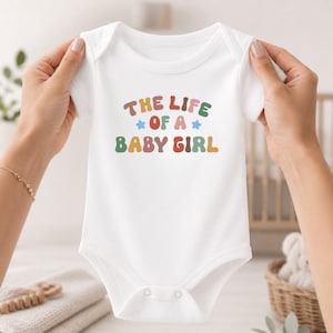 May include: A white short-sleeved baby bodysuit featuring the multicolored text THE LIFE OF A BABY GIRL in a rounded font, accented by two small blue stars. The garment includes a crew neckline and snap closures at the bottom for easy access.