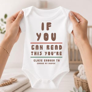 May include: A white short-sleeved baby bodysuit featuring the brown text IF YOU CAN READ THIS YOU'RE CLOSE ENOUGH TO CHANGE MY DIAPER, with a thin red horizontal line above the second line of text and a thin green line below it.
