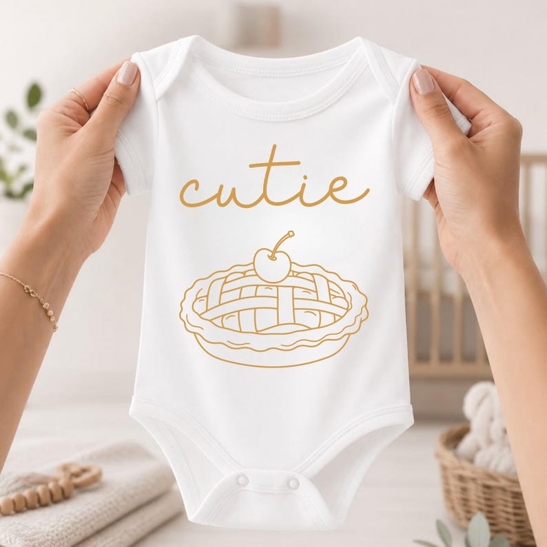 May include: A white baby bodysuit featuring the word cutie in a gold script font above a gold line drawing of a cherry pie. The garment has short sleeves, a round neckline, and a snap closure at the bottom for easy access during diaper changes.