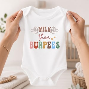 May include: A white short-sleeved baby bodysuit featuring a graphic print with the text "MILK then BURPEES" in colorful letters. The design includes a barbell icon above the text and two small stars, all centered on the front of the garment.