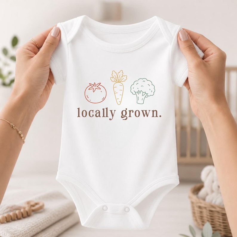 May include: A white short-sleeved baby bodysuit featuring a graphic print of a red tomato, an orange carrot, and a green broccoli floret. Below the vegetables, the text locally grown is printed in a brown, lowercase, sans-serif font.