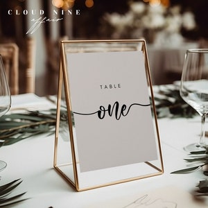 May include: A gold-framed table number sign with a white background and black text. The sign reads "TABLE one".