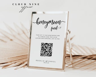 May include: A white sign in a silver frame with the text "honeymoon fund" in black cursive script. The sign also includes a QR code and the text "YOUR GIFT TO OUR HONEYMOON FUND IS A DREAM COME TRUE AS WE EXPLORE NEW HORIZONS, JUST US TWO" and "ROSALIE & DANIEL 05.07.25".