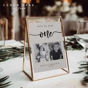 May include: A gold-framed table number with a white background and black text. The text reads "When we were one" with a handwritten font. Two black and white photos of a baby are below the text. The text below the photos reads "Rosalie and Daniel 05.07.25".