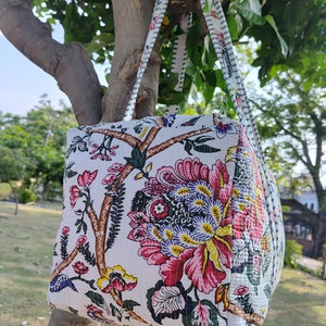 May include: A white tote bag with floral print featuring pink, red, yellow, and green flowers. The bag has two white straps with black trim.