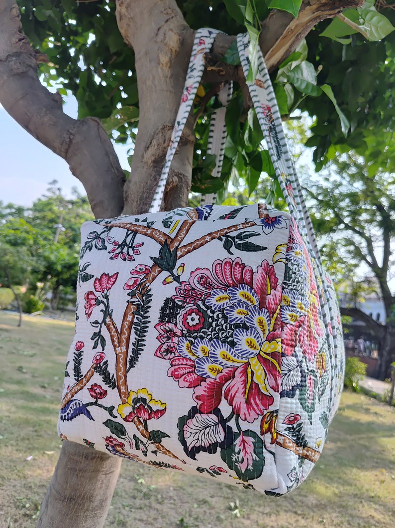 May include: A white tote bag with floral print featuring pink, red, yellow, and green flowers. The bag has two white straps with black trim.