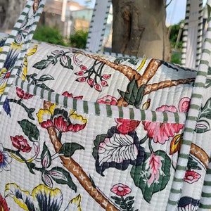May include: A white quilted tote bag with a floral print and green and white striped straps. The bag has a brown branch design and pink, red, and yellow flowers.