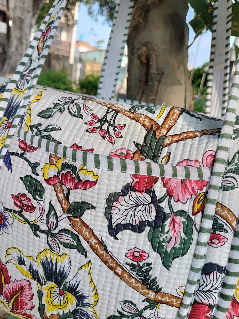 May include: A white quilted tote bag with a floral print and green and white striped straps. The bag has a brown branch design and pink, red, and yellow flowers.