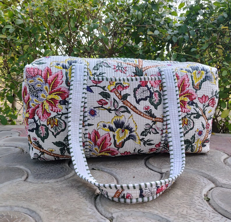 May include: White cotton travel bag with a floral print in shades of pink, yellow, green, and blue. The bag has two white straps with green trim.