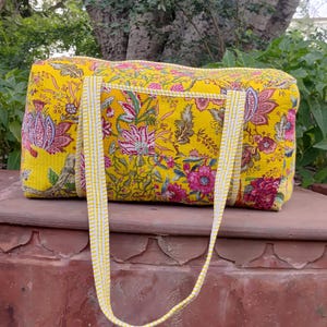 Puede incluir: Una bolsa de viaje amarilla con estampado floral y asas a rayas blancas. La bolsa presenta un diseño vibrante con elementos florales rosas, rojos y verdes. La bolsa es rectangular y parece estar hecha de tela acolchada.