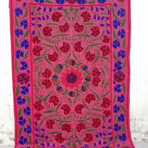 Könnte beinhalten: Eine rosa Baumwoll-Wandbehang mit einem floralen Muster, das in roter, grüner und blauer Farbe gestickt ist. Der Wandbehang zeigt ein zentrales Motiv einer stilisierten Blume, die von kleineren Blumen und Blättern umgeben ist.