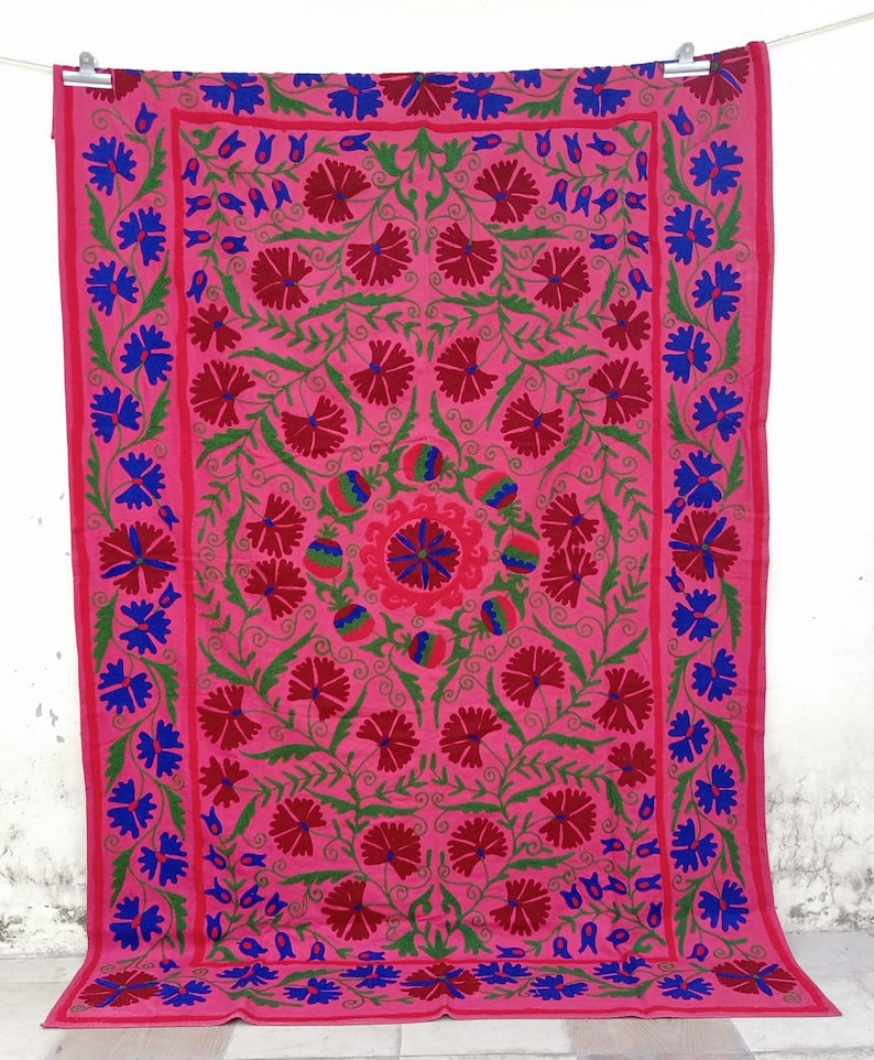 May include: A pink cotton tapestry with a floral design embroidered in red, green, and blue thread. The tapestry features a central motif of a stylised flower surrounded by smaller flowers and leaves.