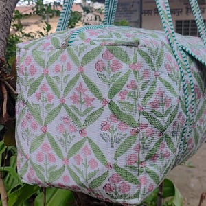 May include: A white duffel bag with a floral pattern in pink and green. The bag has green and white striped handles and trim. The floral pattern is a repeating design of flowers and leaves.