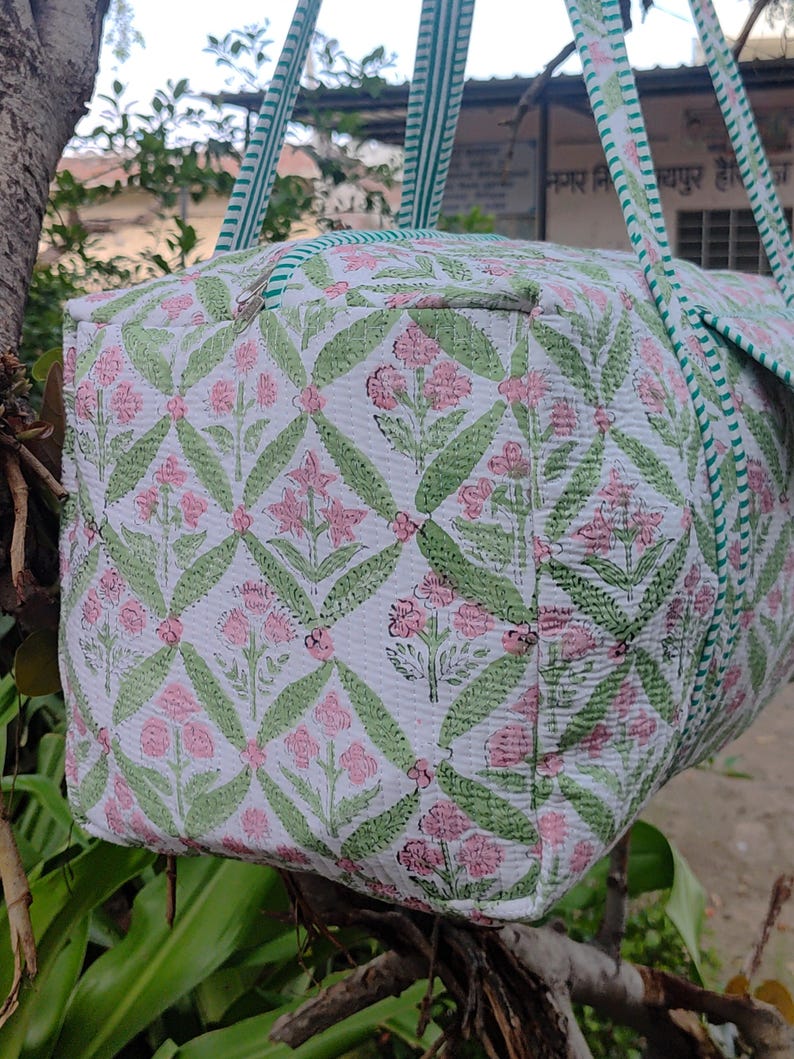 May include: A white duffel bag with a floral pattern in pink and green. The bag has green and white striped handles and trim. The floral pattern is a repeating design of flowers and leaves.