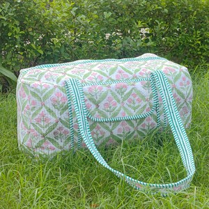 May include: A white and pink floral patterned duffel bag with green trim and handles. The bag is rectangular with a pocket on the front and is sitting on green grass.