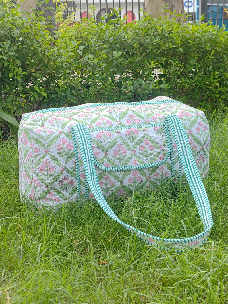 May include: A white and pink floral patterned duffel bag with green trim and handles. The bag is rectangular with a pocket on the front and is sitting on green grass.