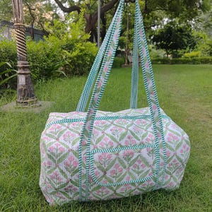 May include: A white and pink floral patterned tote bag with green trim and handles. The bag has a rectangular shape with a pocket on the front. The handles are green and white striped. The bag is held up by a hand.
