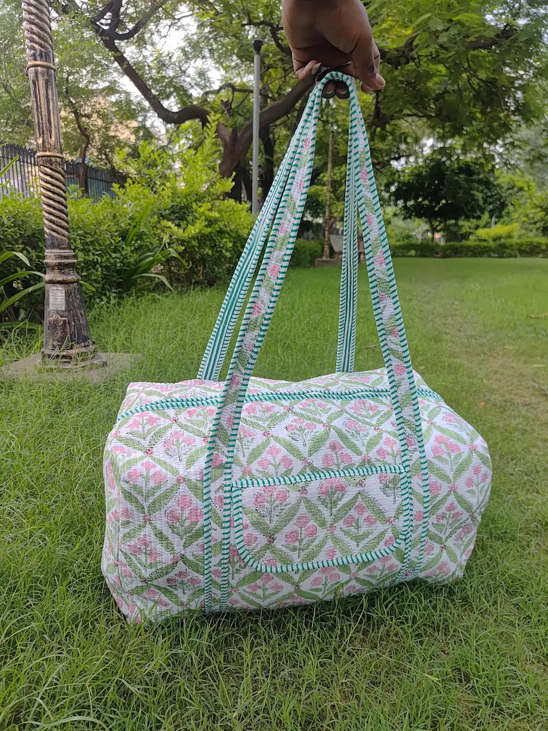 May include: A white and pink floral patterned tote bag with green trim and handles. The bag has a rectangular shape with a pocket on the front. The handles are green and white striped. The bag is held up by a hand.