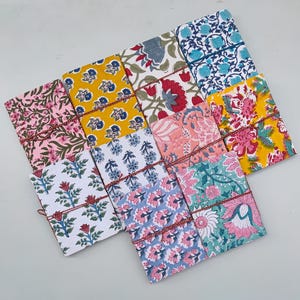 Könnte beinhalten: Ein Set aus zehn bunten, mit Stoff bezogenen Notizbüchern mit Blumenmustern. Die Notizbücher sind mit roter Schnur gebunden und haben eine Vielzahl von Farben, darunter Gelb, Rosa, Blau und Grün.
