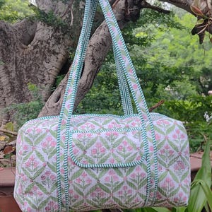 May include: A quilted duffel bag with a floral pattern in pink and green on a white background. The bag has matching straps and a green trim. The bag is hanging from a tree branch.