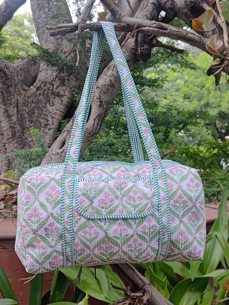 May include: A quilted duffel bag with a floral pattern in pink and green on a white background. The bag has matching straps and a green trim. The bag is hanging from a tree branch.