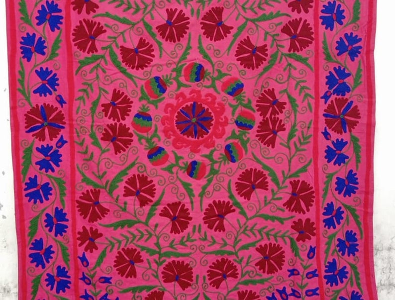 May include: A pink fabric with intricate embroidery featuring red and blue floral designs. The fabric has a central motif with a circular pattern and is surrounded by repeating floral patterns.
